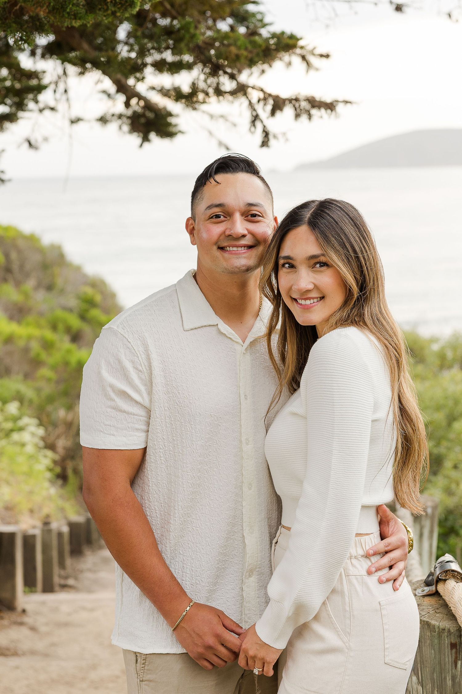 A couple in matching white outfits poses together outdoors with trees in the background.
