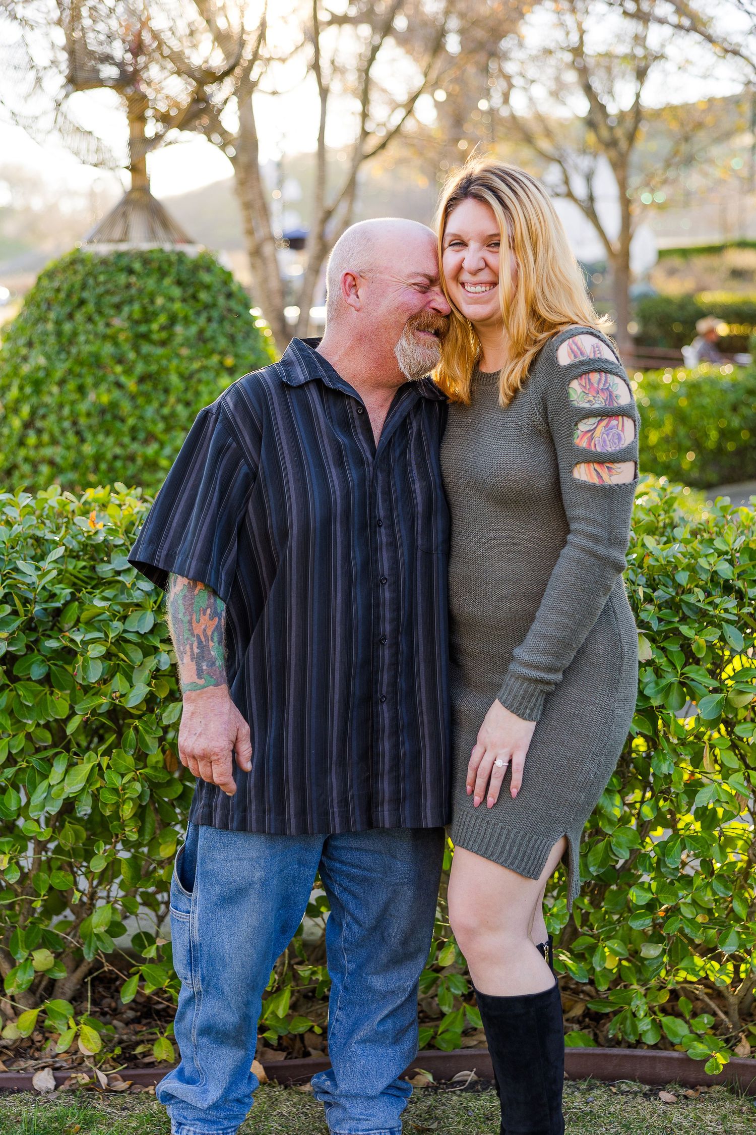 A couple embraces while standing in front of lush green hedges during golden hour.