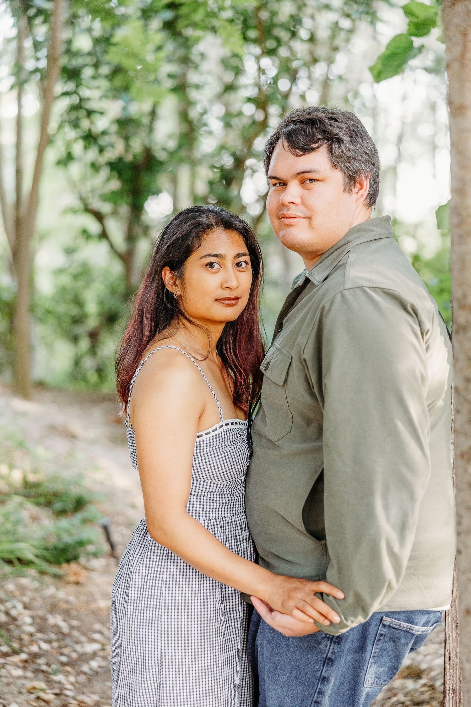 A couple embraces while standing among trees during an outdoor photoshoot in natural lighting.