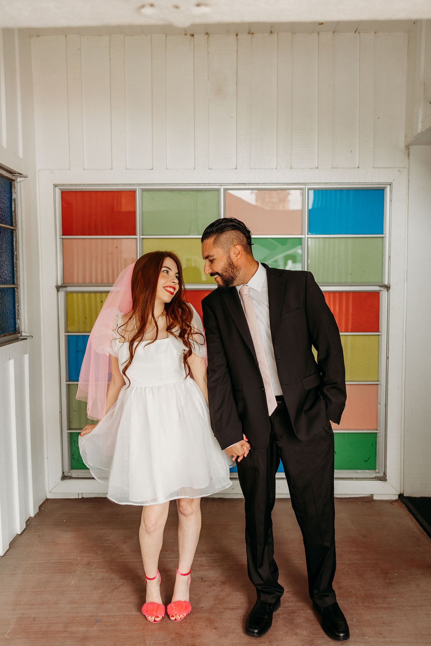 Couple in formal attire laughing together against a colorful paneled wall with pink and blue squares.