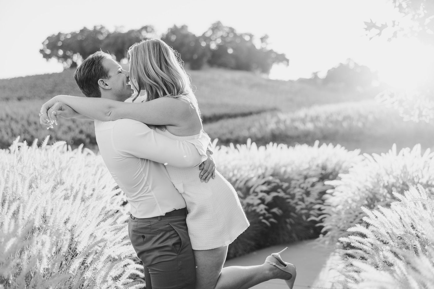 A couple embraces in a romantic moment surrounded by a sunlit field of tall grass and flowers.