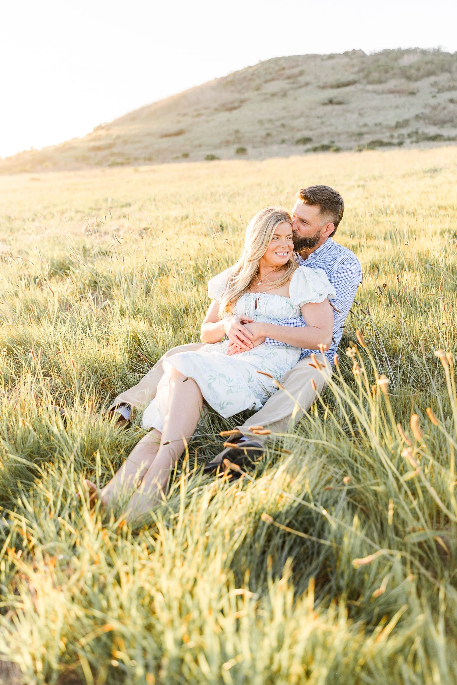 A romantic couple embraces in a sunlit golden field of tall grass at sunset with rolling hills in the background.