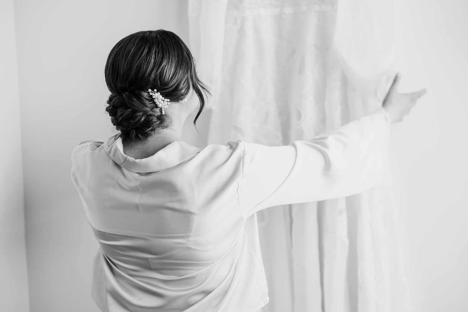 Black and white photo of person in white dress reaching toward window curtains.