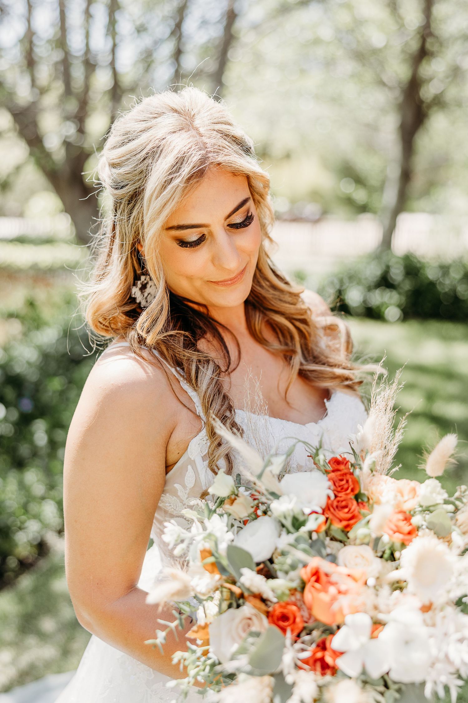 A bride in a white wedding dress holds a beautiful coral and white bouquet while standing outdoors in nature.