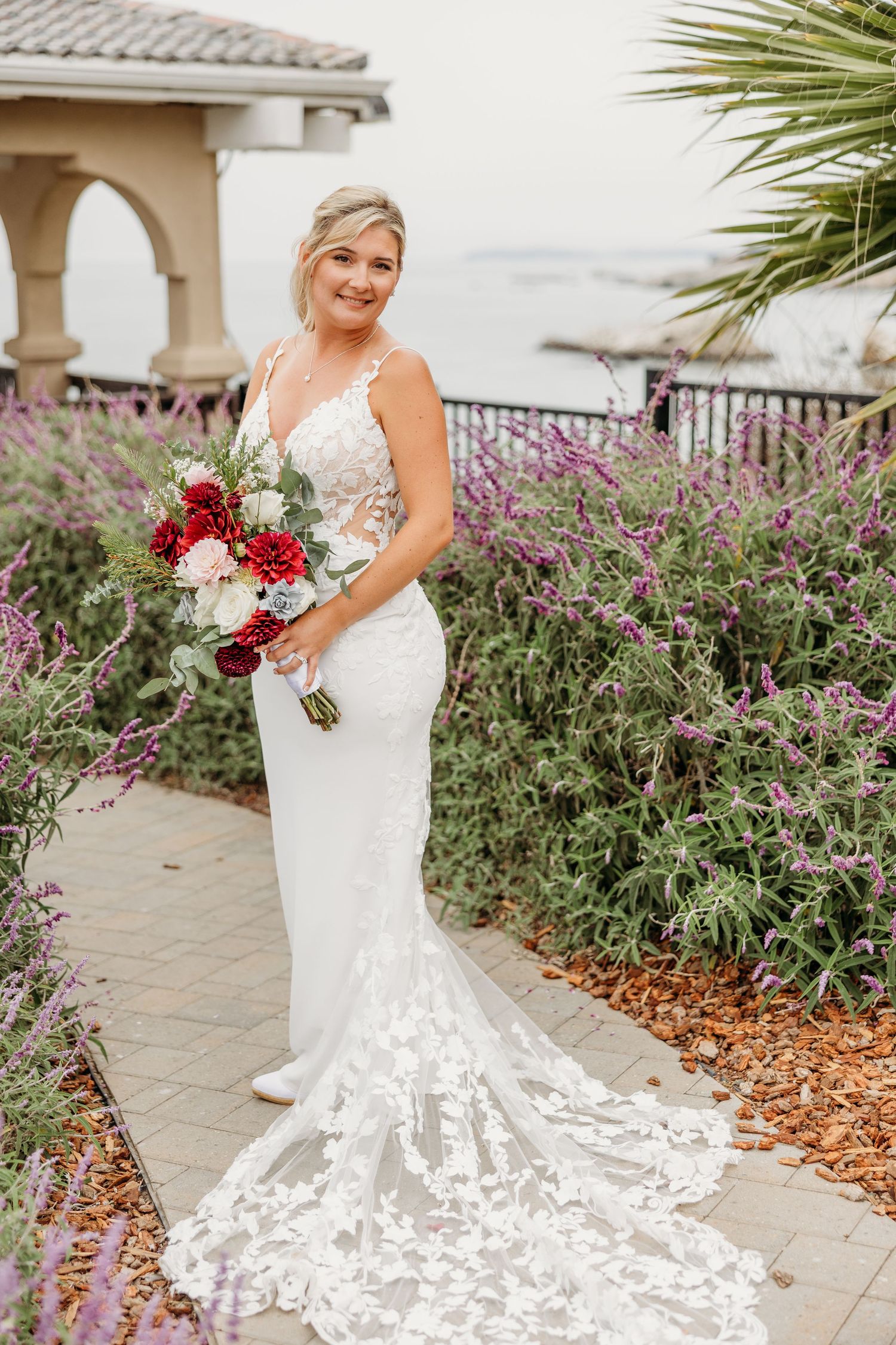Elegant bridal portrait featuring a fitted lace gown and red and white bouquet by the ocean.
