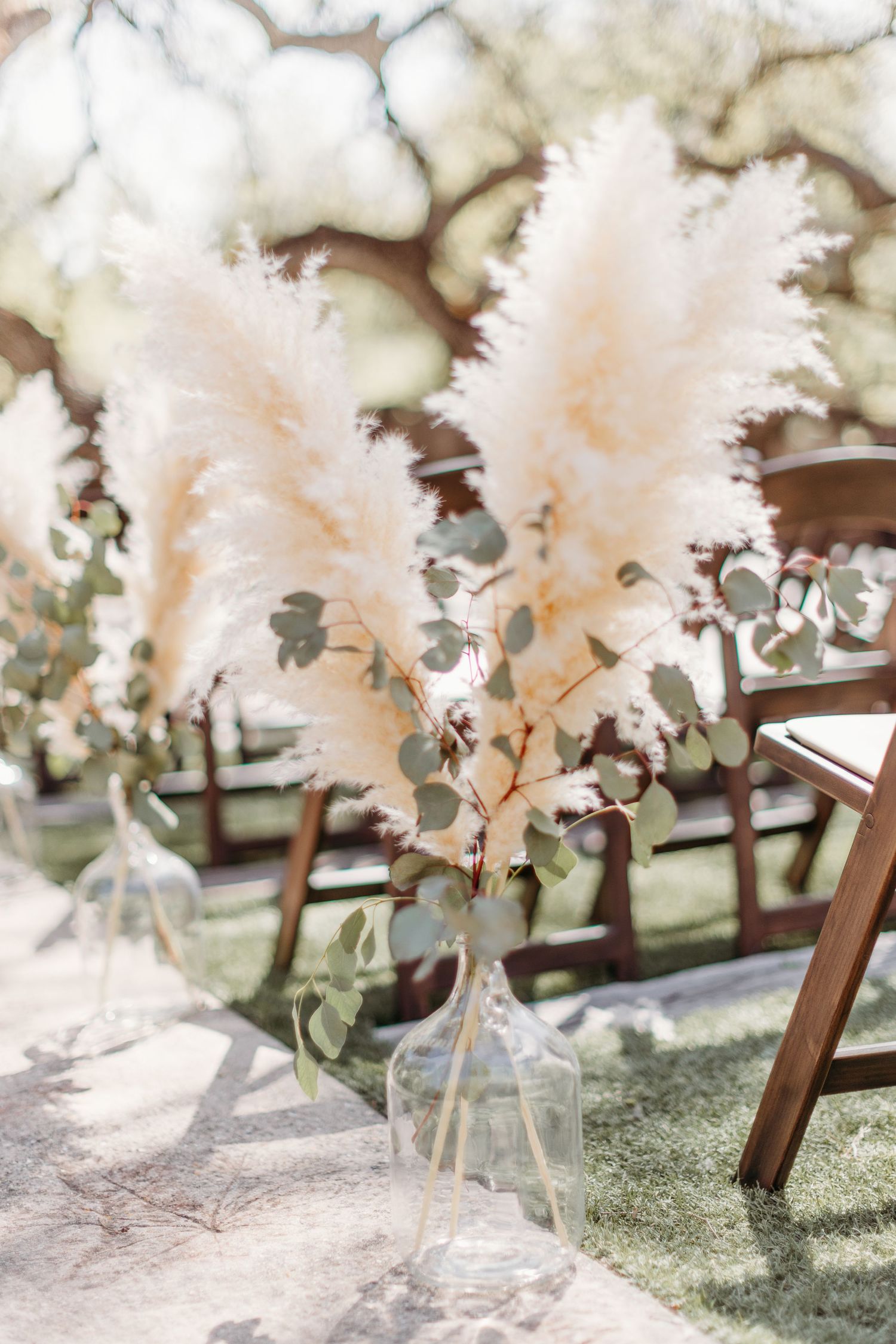 Soft pink pampas grass and eucalyptus arrangement in clear glass vase beside wooden chair for outdoor wedding decor.