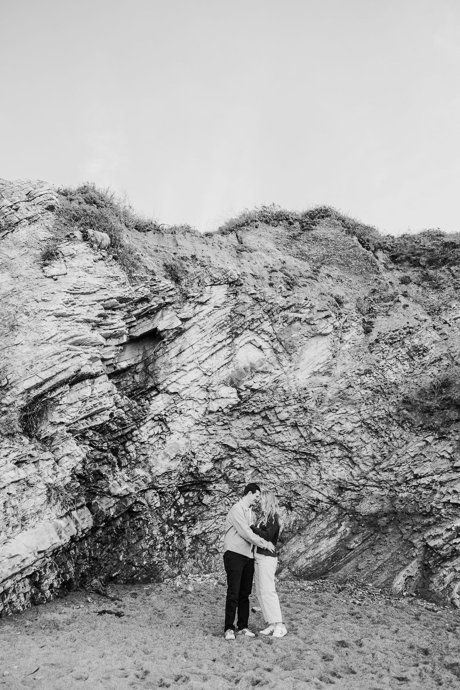 Two people embrace in front of dramatic rocky cliffs in black and white.