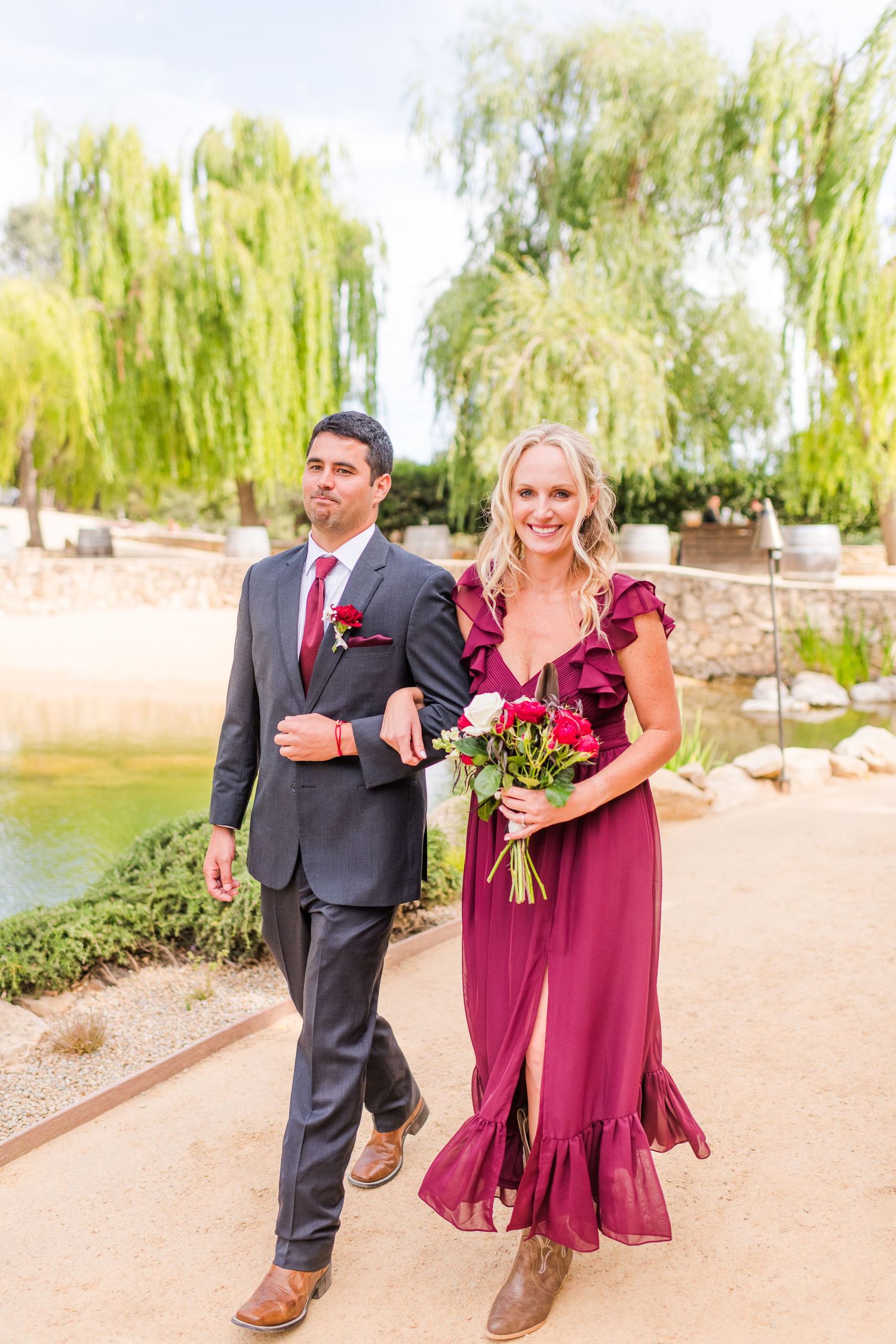 A couple in formal attire walking arm in arm along a garden pathway.