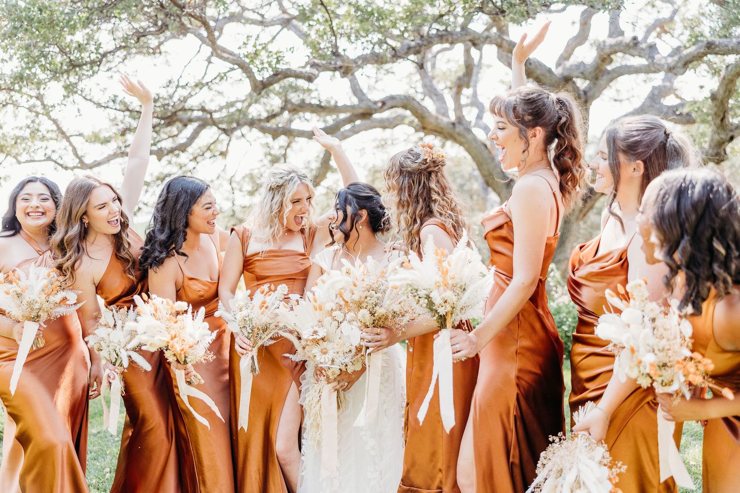 Bridesmaids in rust-colored silk dresses holding white bouquets stand together beneath sprawling trees.