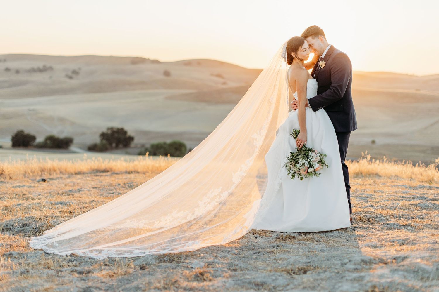 Long flowing wedding veil catches golden sunlight across rolling hills during sunset photography session.