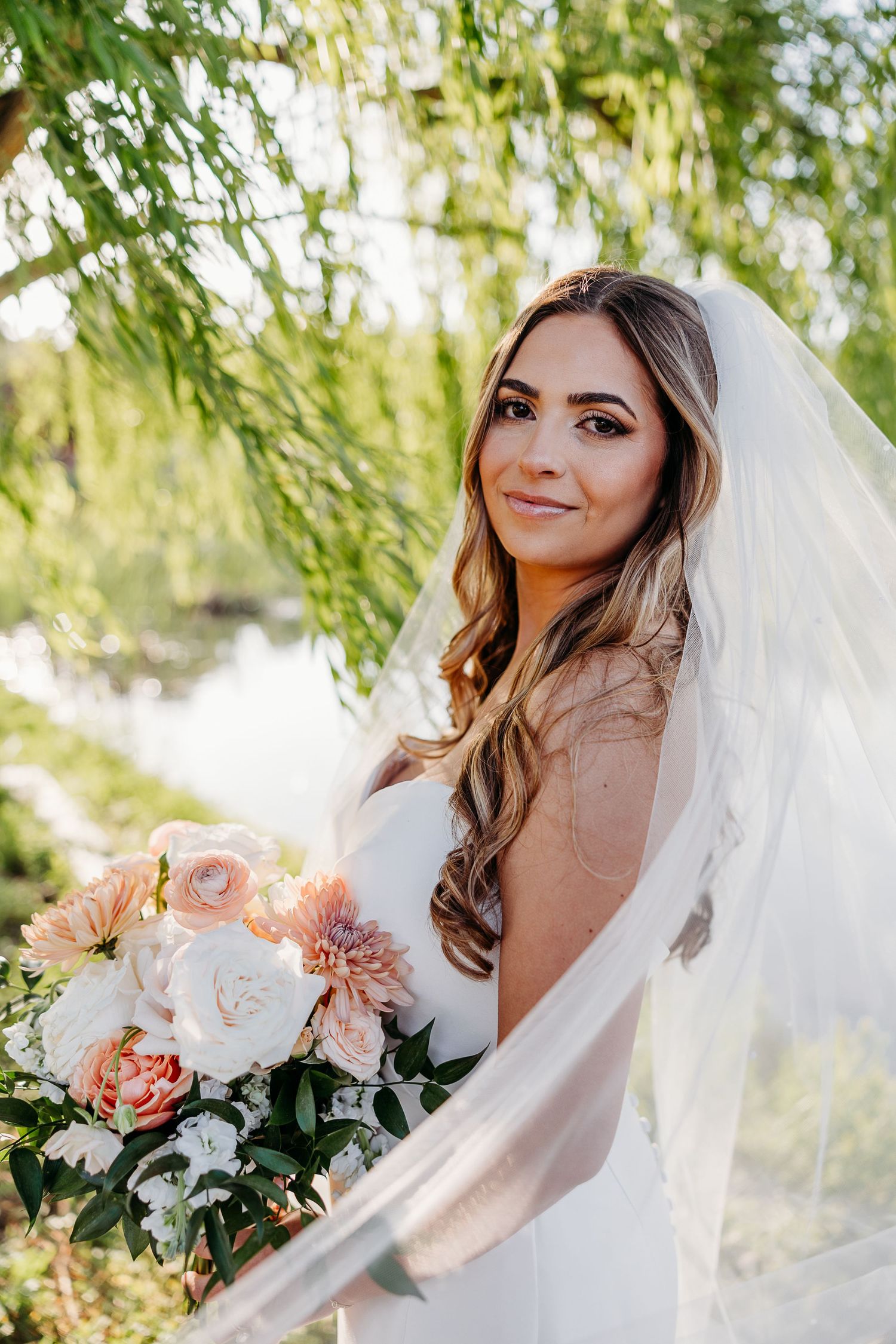Elegant bride in white gown holds peach and cream bouquet under willow tree branches.