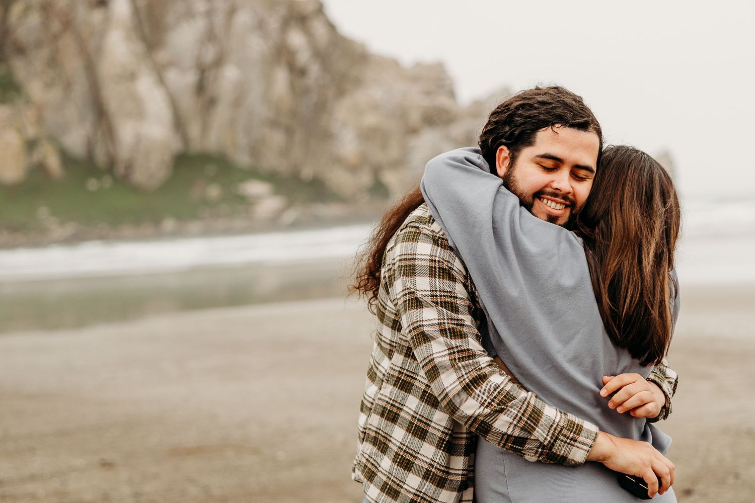 A couple embraces in a warm hug on a misty beach with rocky cliffs in the background.