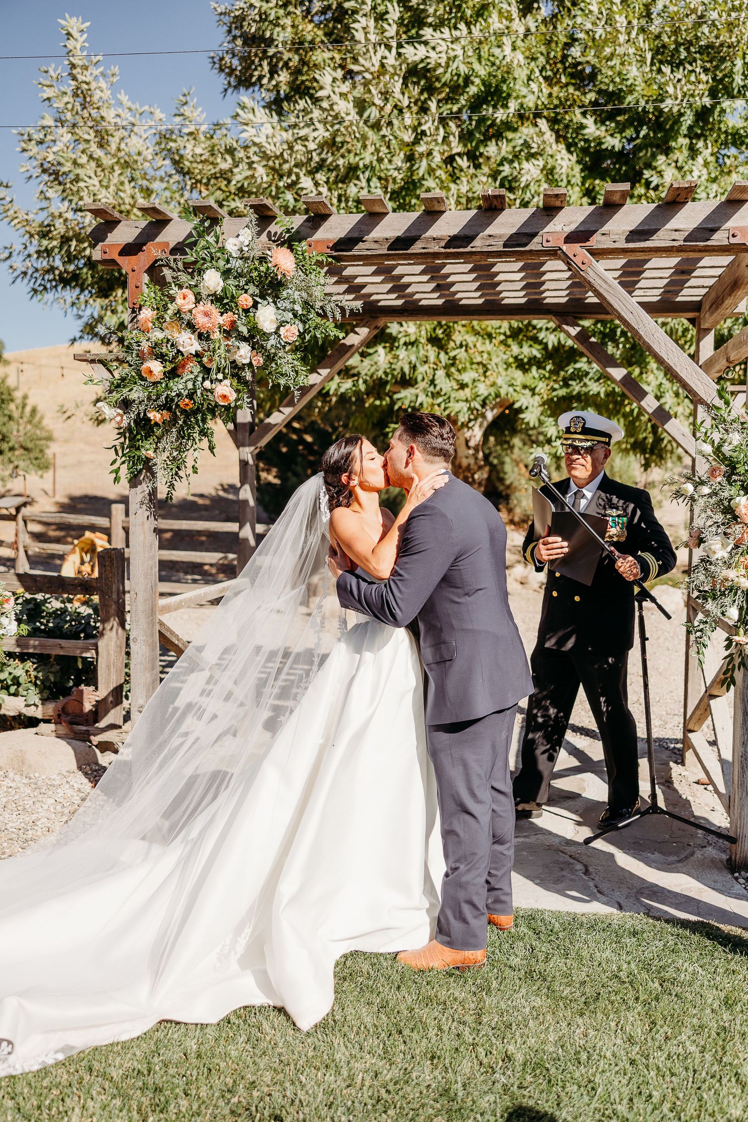 A romantic wedding ceremony takes place under a wooden pergola with flowing white dress and veil in an outdoor garden setting.