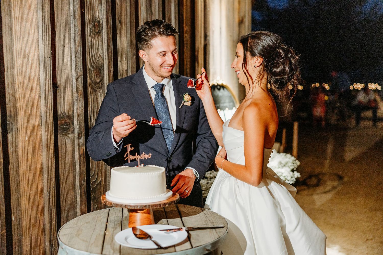 Newlyweds share a laugh while cutting their wedding cake on rustic barrel table.