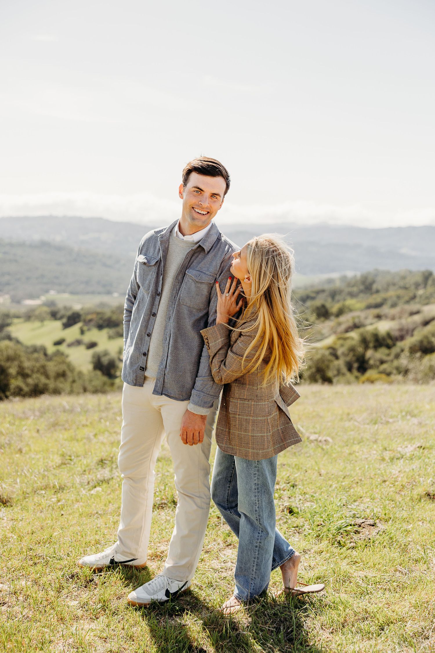A couple shares a joyful moment together in a scenic meadow with rolling hills in the background.