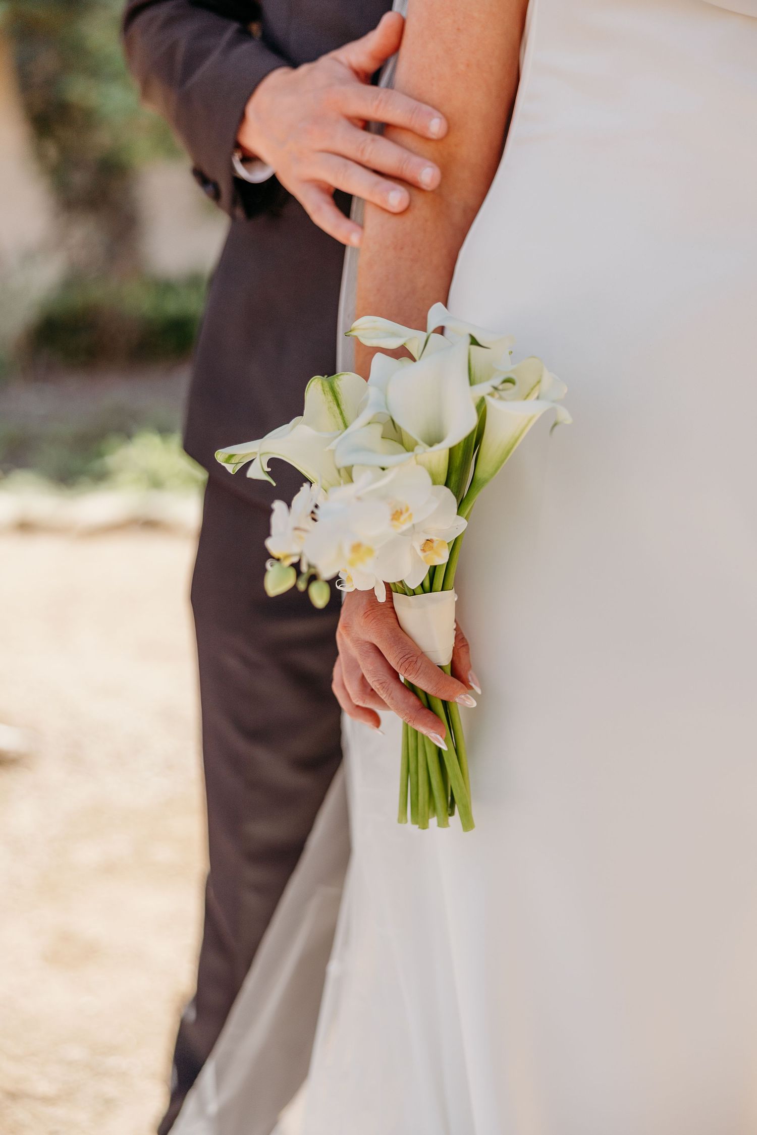 A close-up of a white wedding bouquet with lilies being held against a white dress and dark suit.