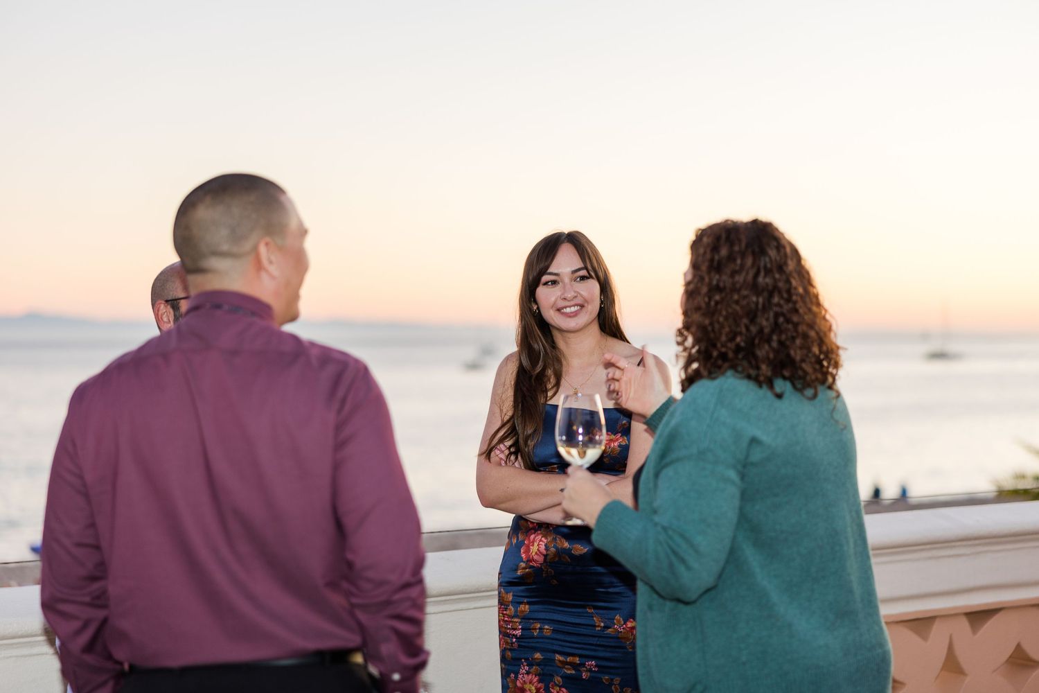 Three people enjoying conversation and wine on a waterfront patio during sunset.