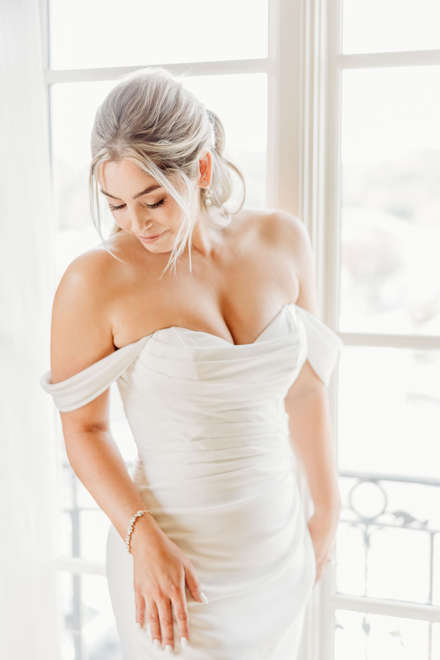 A bride in an elegant off-shoulder white wedding gown poses by a window.