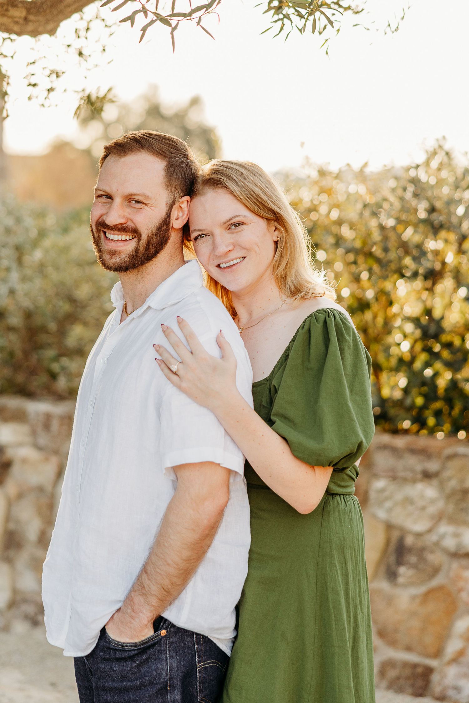 Happy couple laughing and embracing outdoors in front of rustic stone wall during sunset photoshoot.