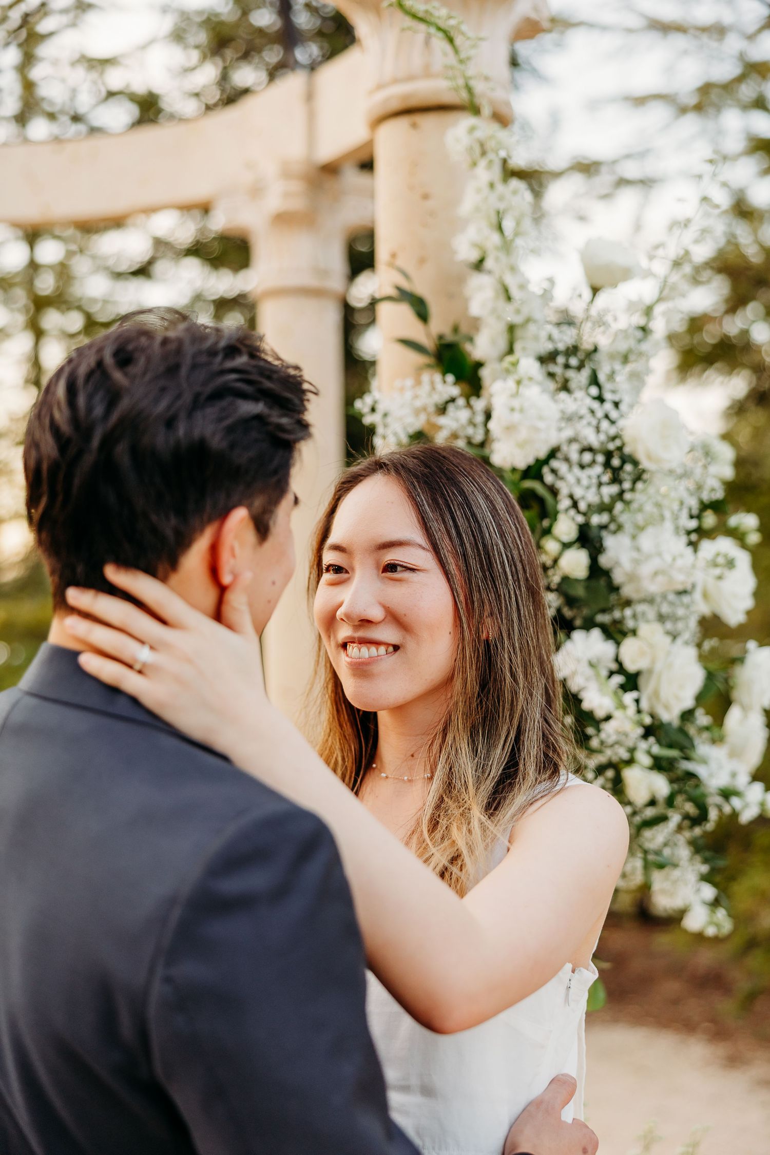 Romantic couple embracing and laughing under a white floral arch during golden hour.
