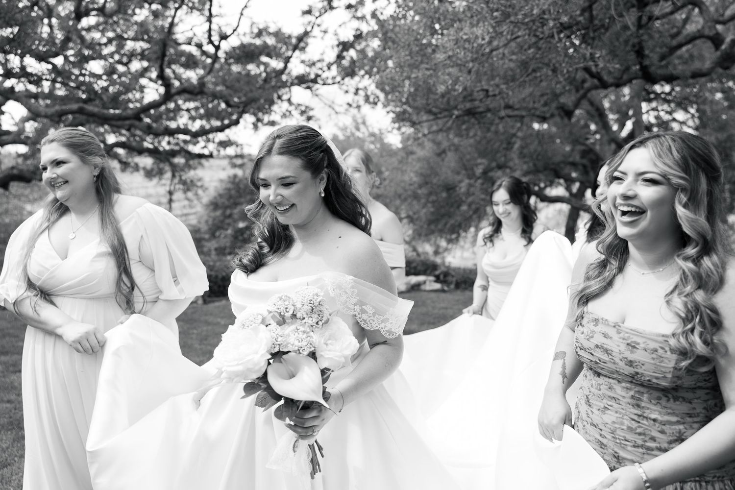 Bride walks with bridesmaids outdoors in black and white wedding photo, holding bouquet.