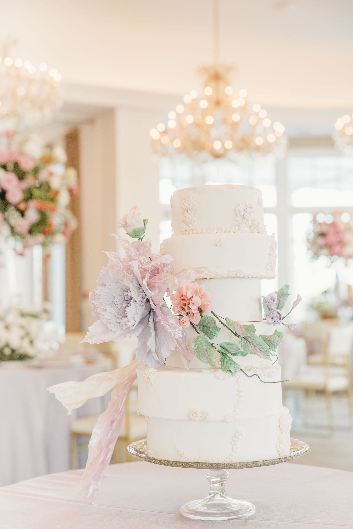 Elegant white wedding cake decorated with delicate pink flowers and greenery sits on display in a bright venue.