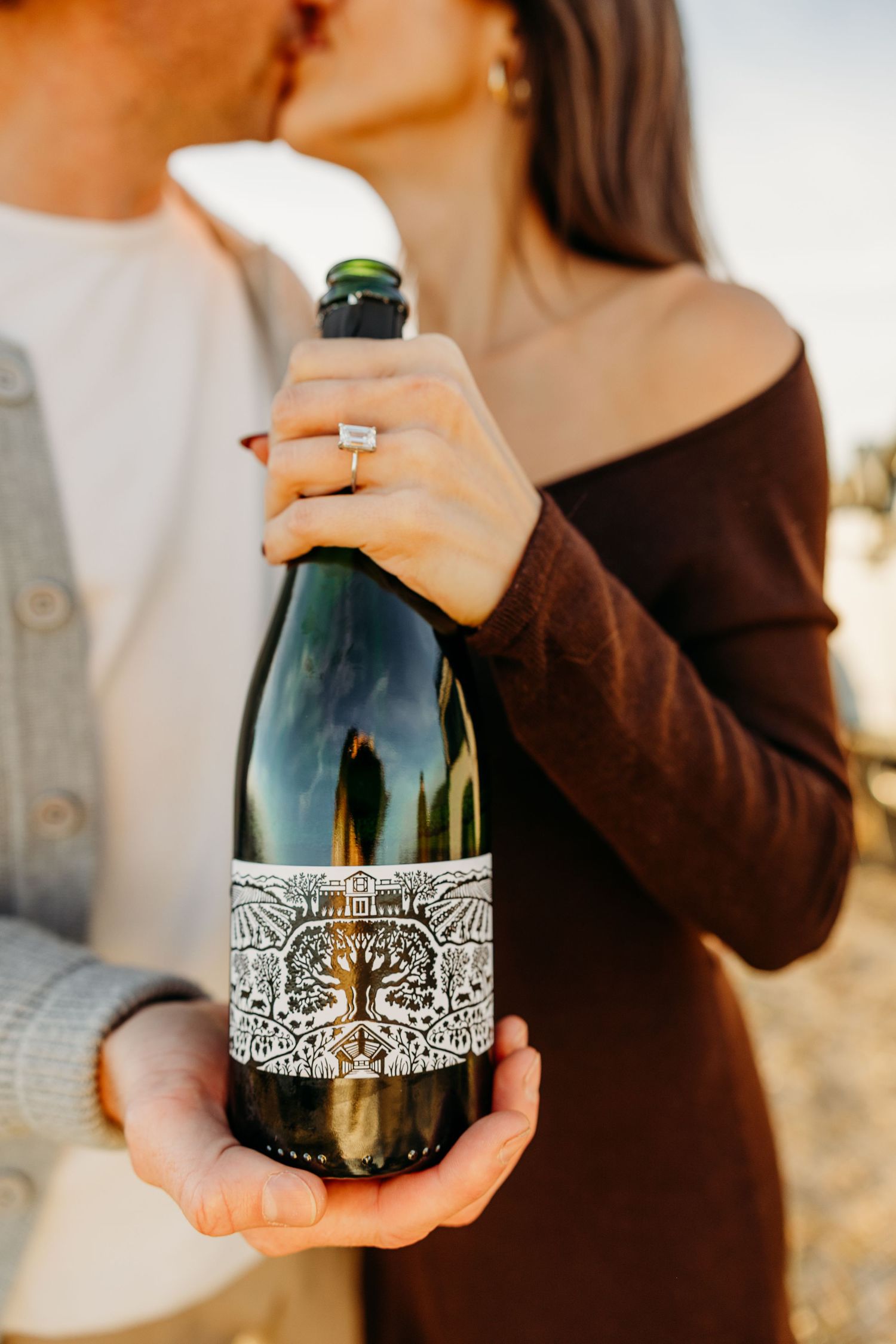 Hands holding a dark glass bottle with an ornate white lace pattern design against a blurred background.