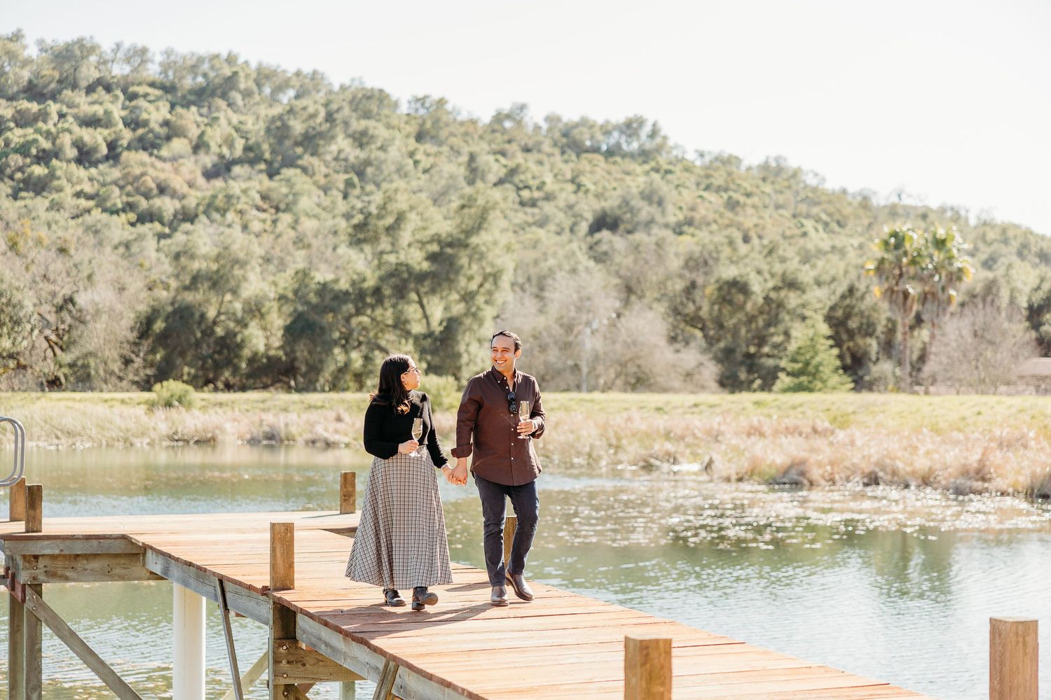 A couple walks together on a wooden dock overlooking a serene lake and hills.