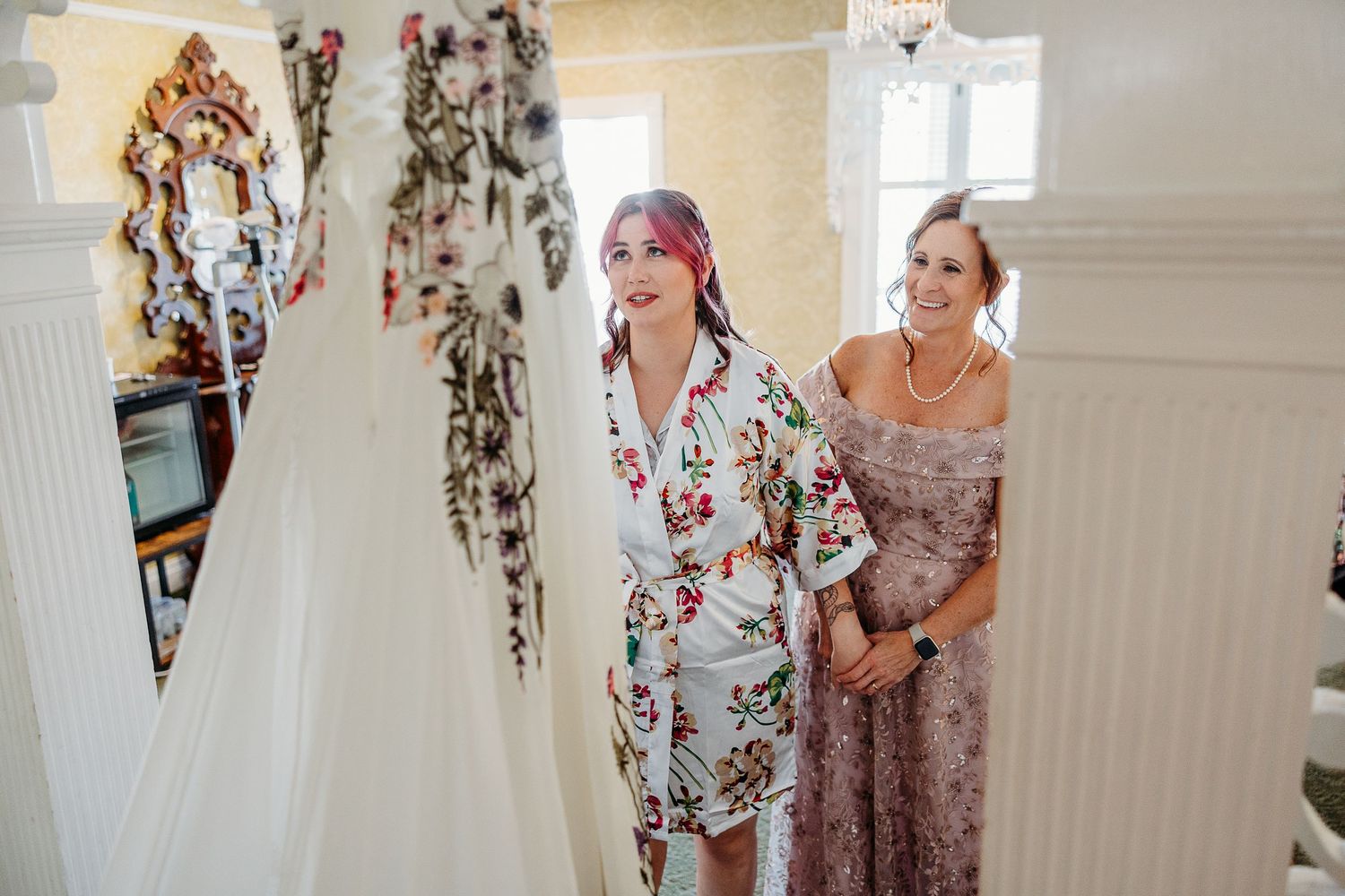 Two women in floral dresses stand near a white wedding gown hanging on display in a bright room.