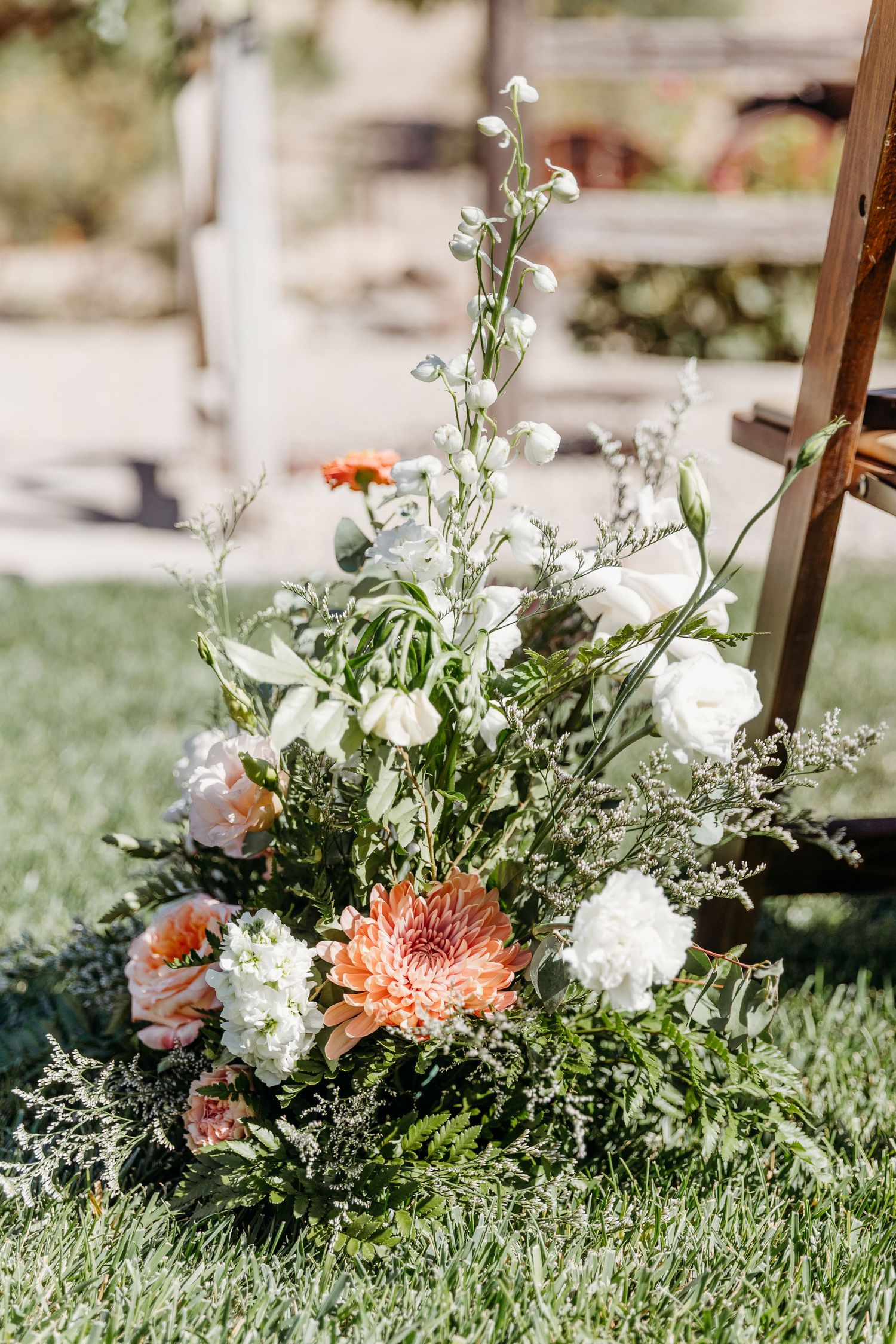 Delicate floral arrangements with coral and white blooms line a grassy outdoor wedding aisle.