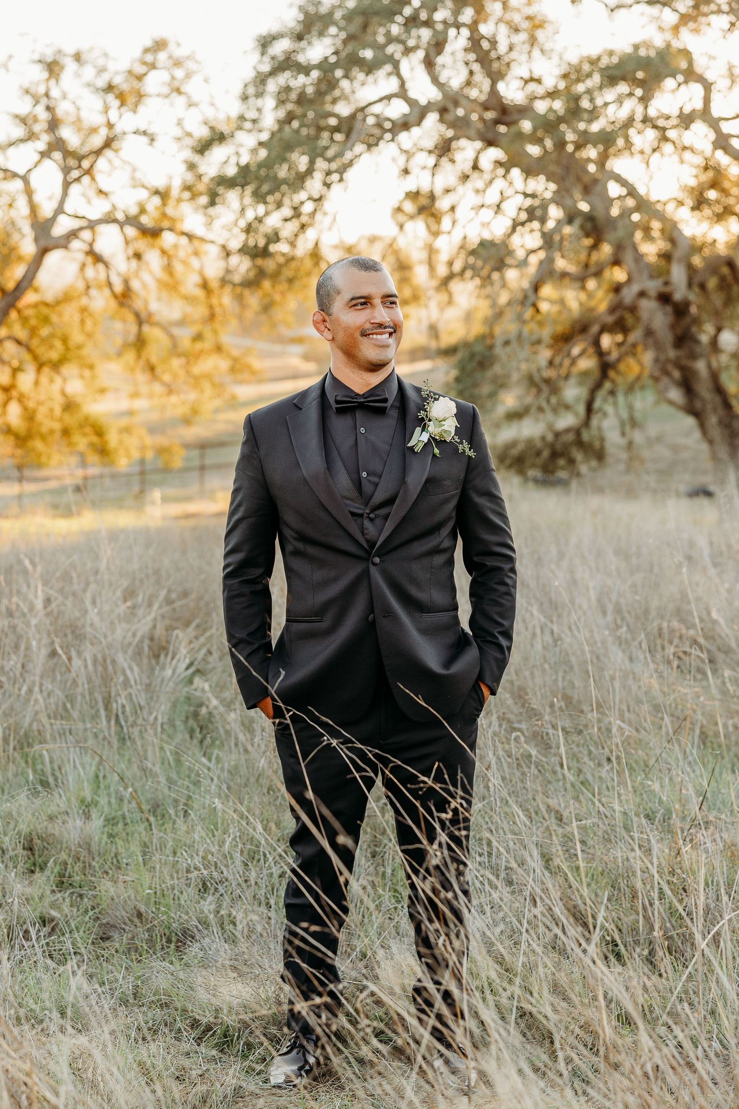 Groom in classic black tuxedo poses in tall grass during sunset wedding photoshoot.
