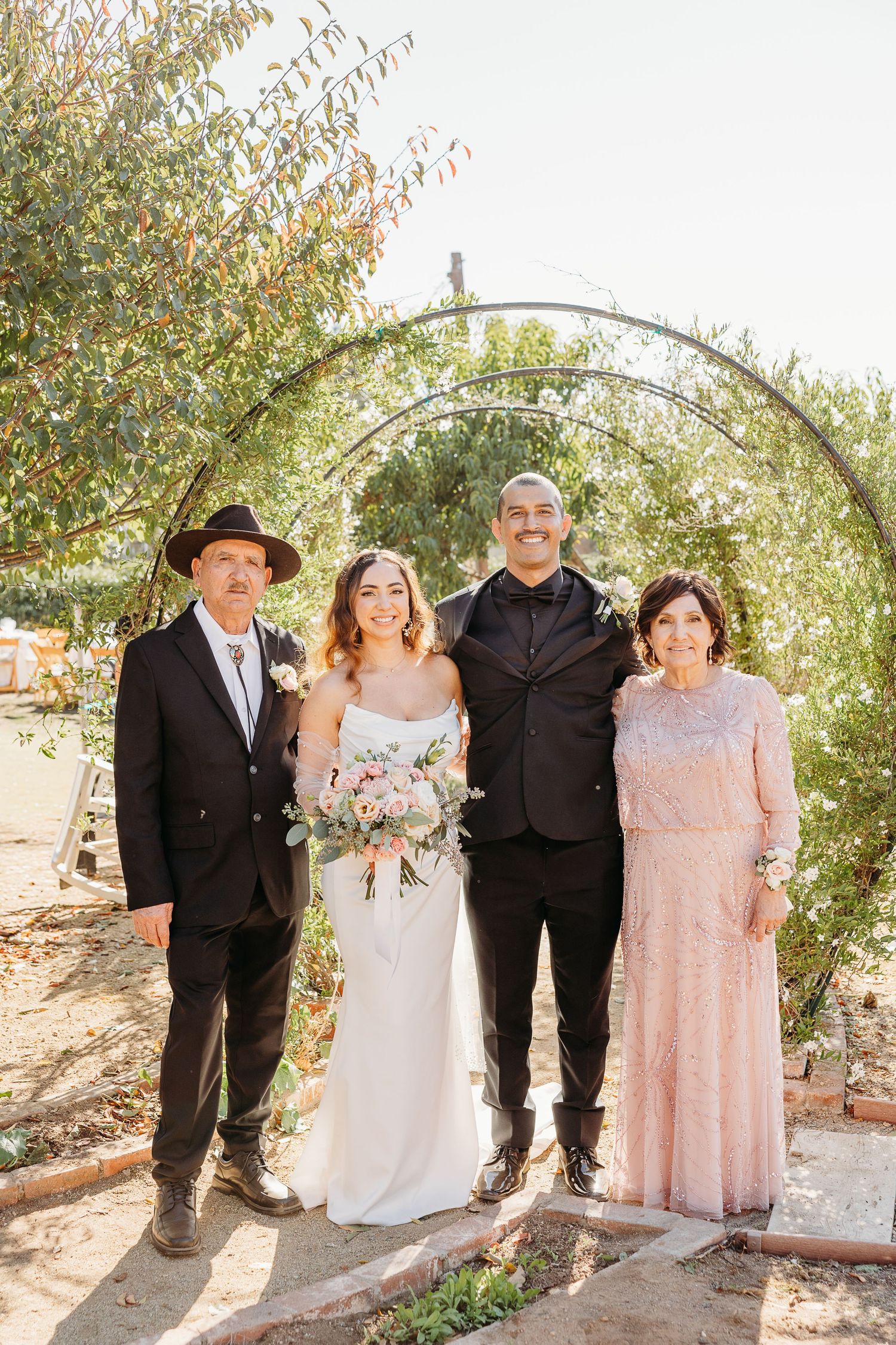 Wedding group photo under an arch with guests wearing pink and black formal attire.