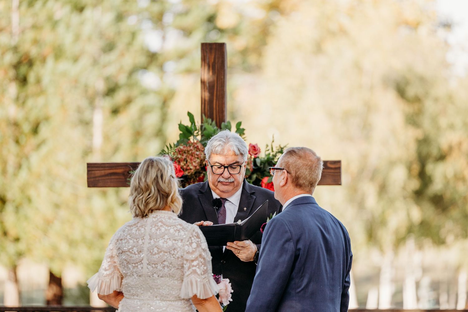 Wedding ceremony in progress with officiant and couple in front of wooden cross outdoors.
