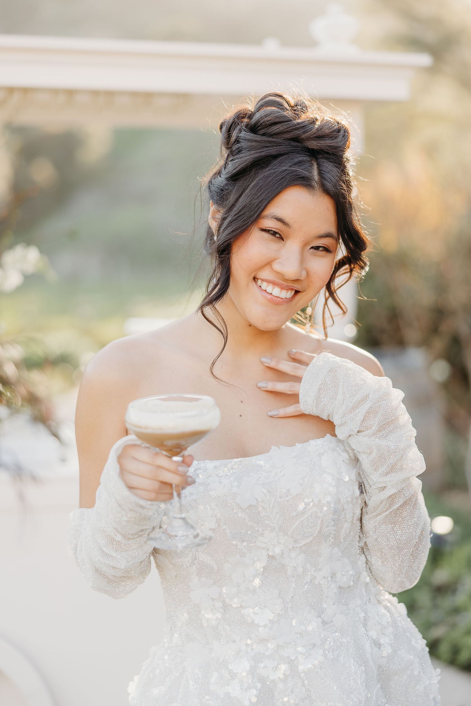 Bride in sparkling long-sleeve wedding gown enjoying celebratory drink outdoors.