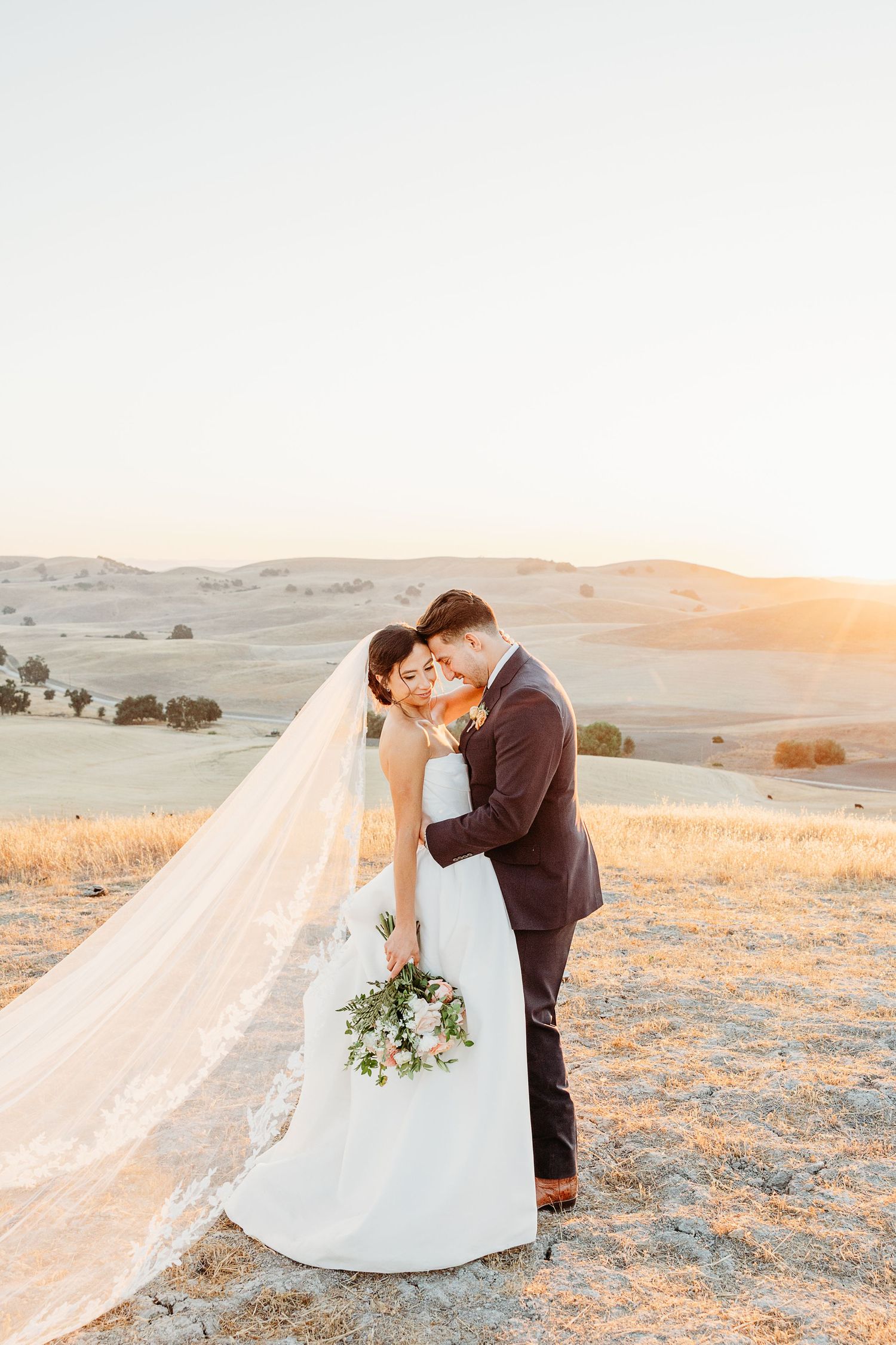 Bride and groom share romantic sunset moment in desert landscape with flowing veil and mountains in background.