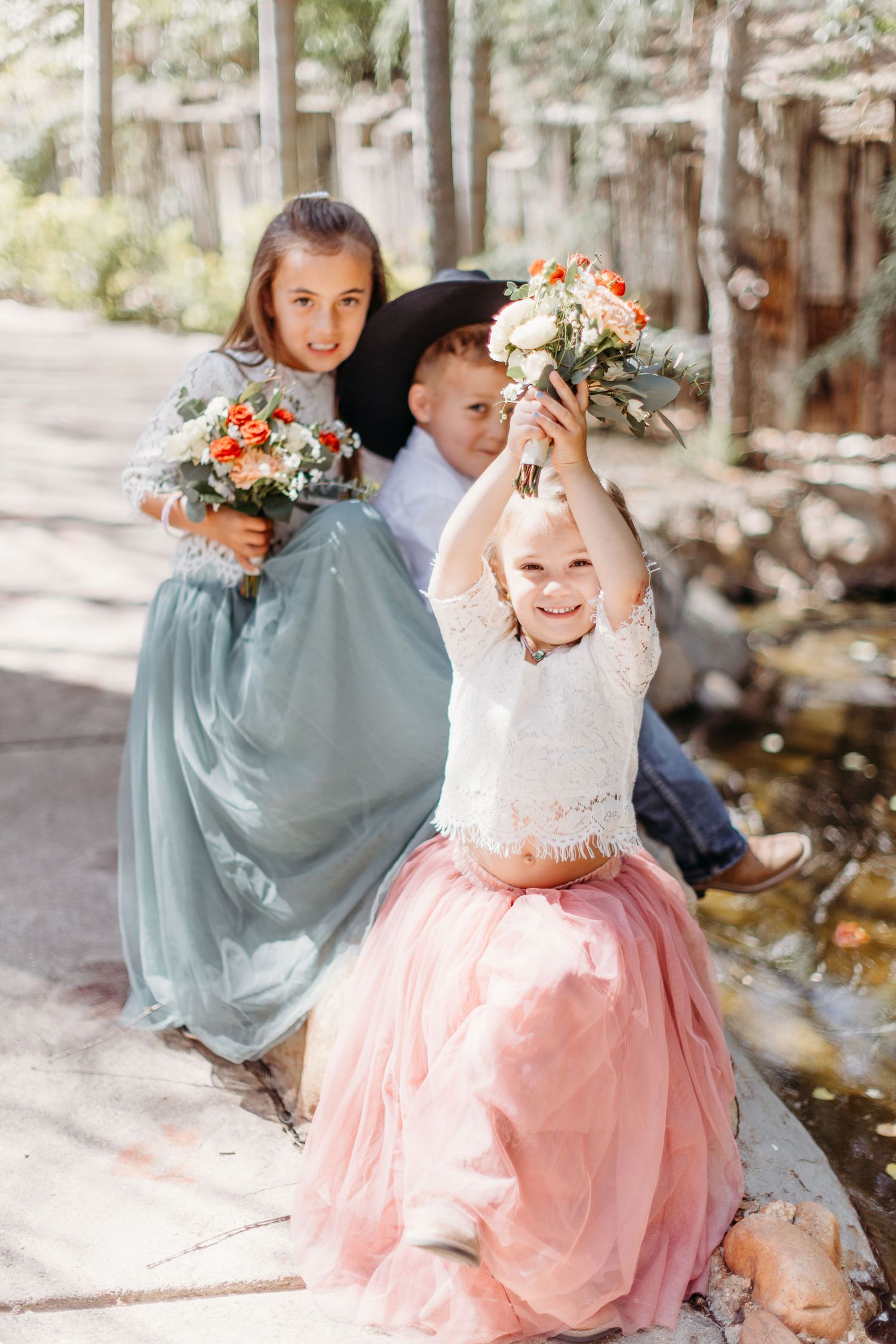 Three children in formal attire hold bouquets while playing and laughing on a wooded path.