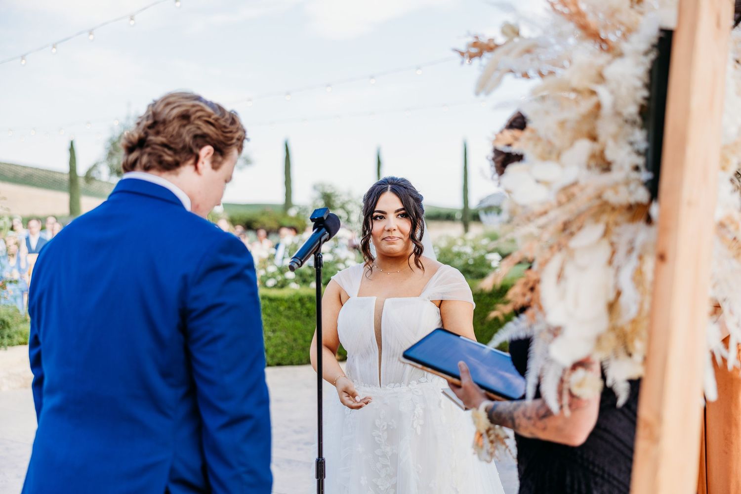 Couple exchanges vows during outdoor vineyard wedding ceremony surrounded by floral arrangements.