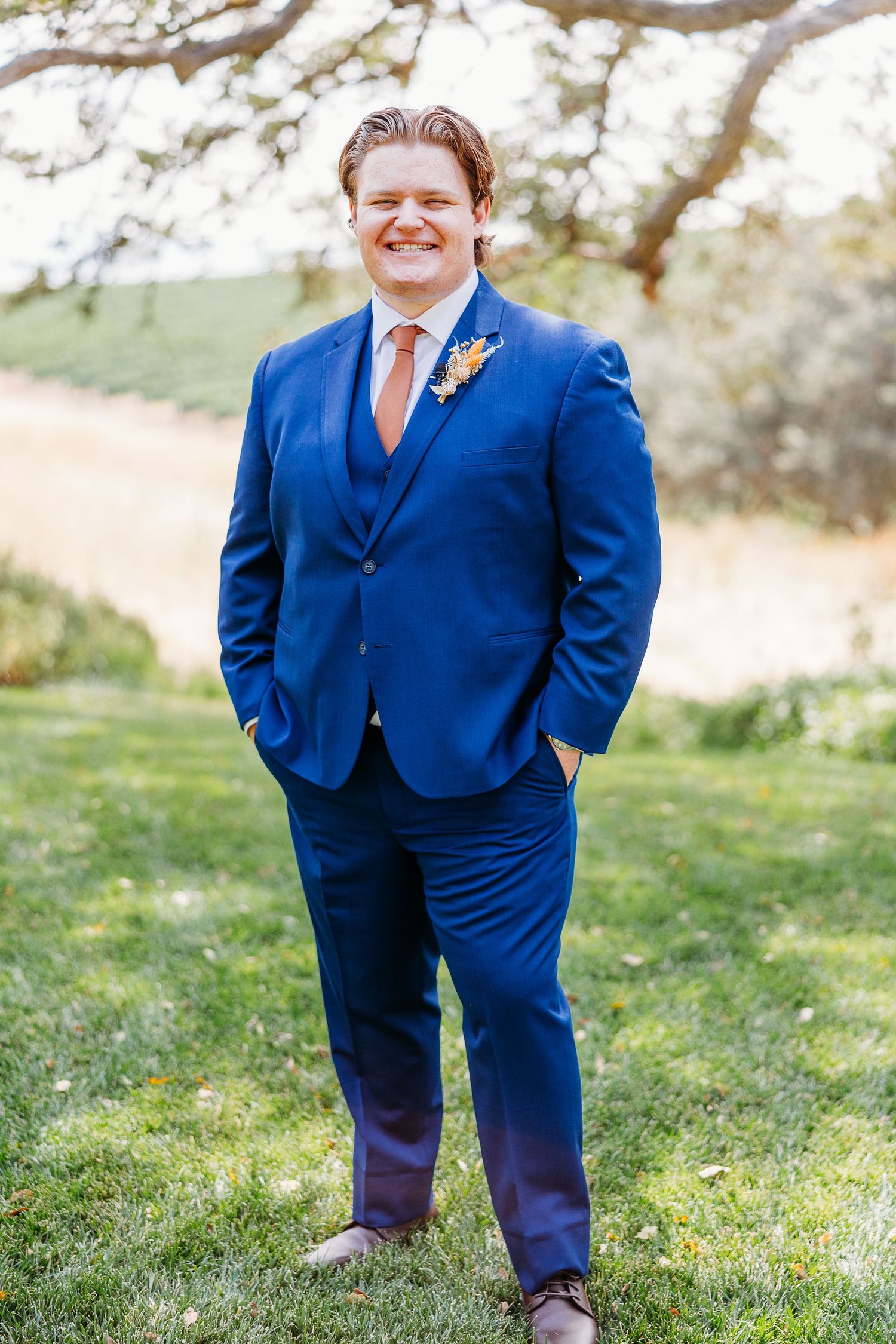 A person in a bright blue suit smiles while standing on grass with trees in background.
