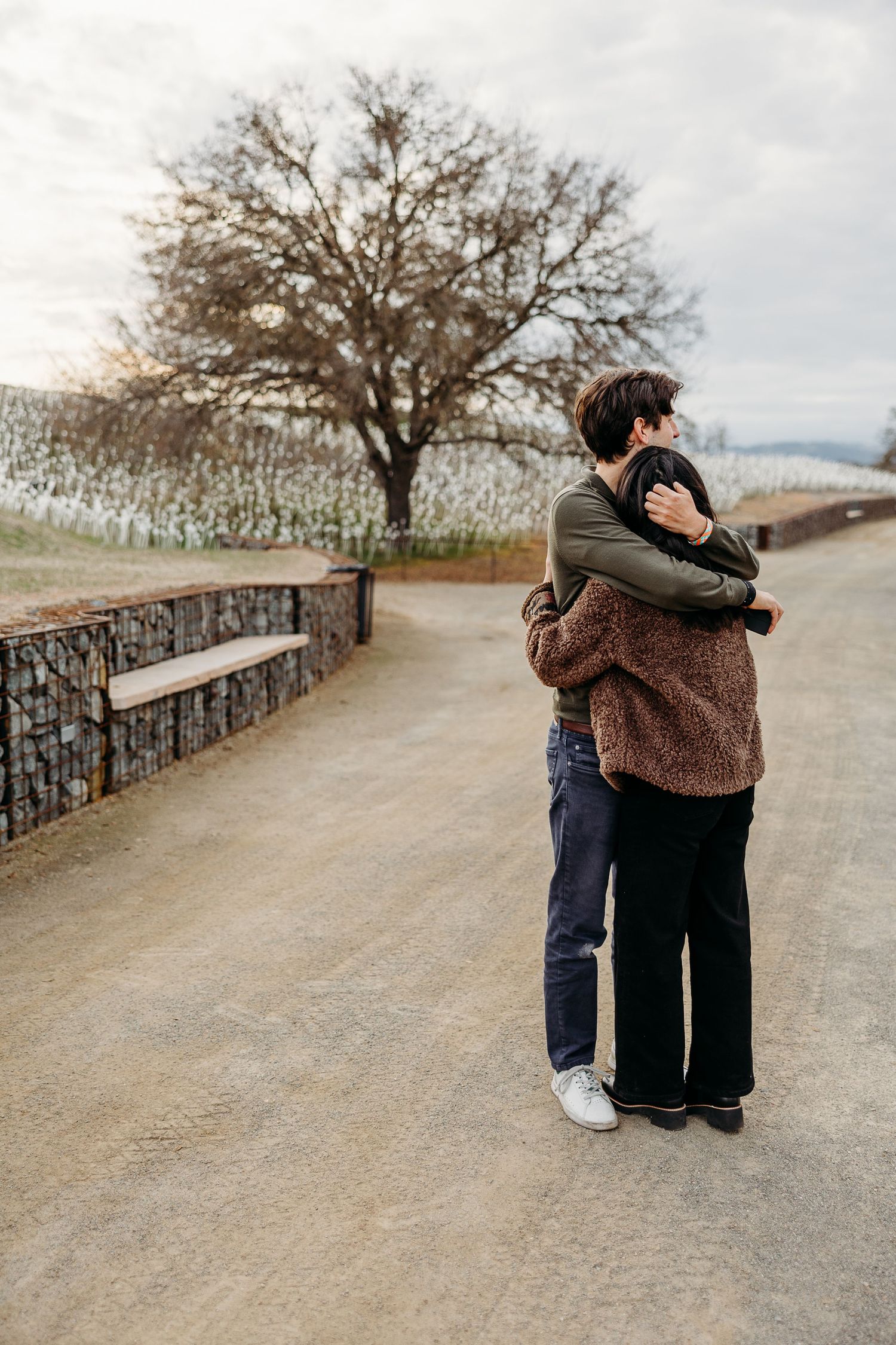Two people embracing on a path with bare trees and cloudy sky in the background.