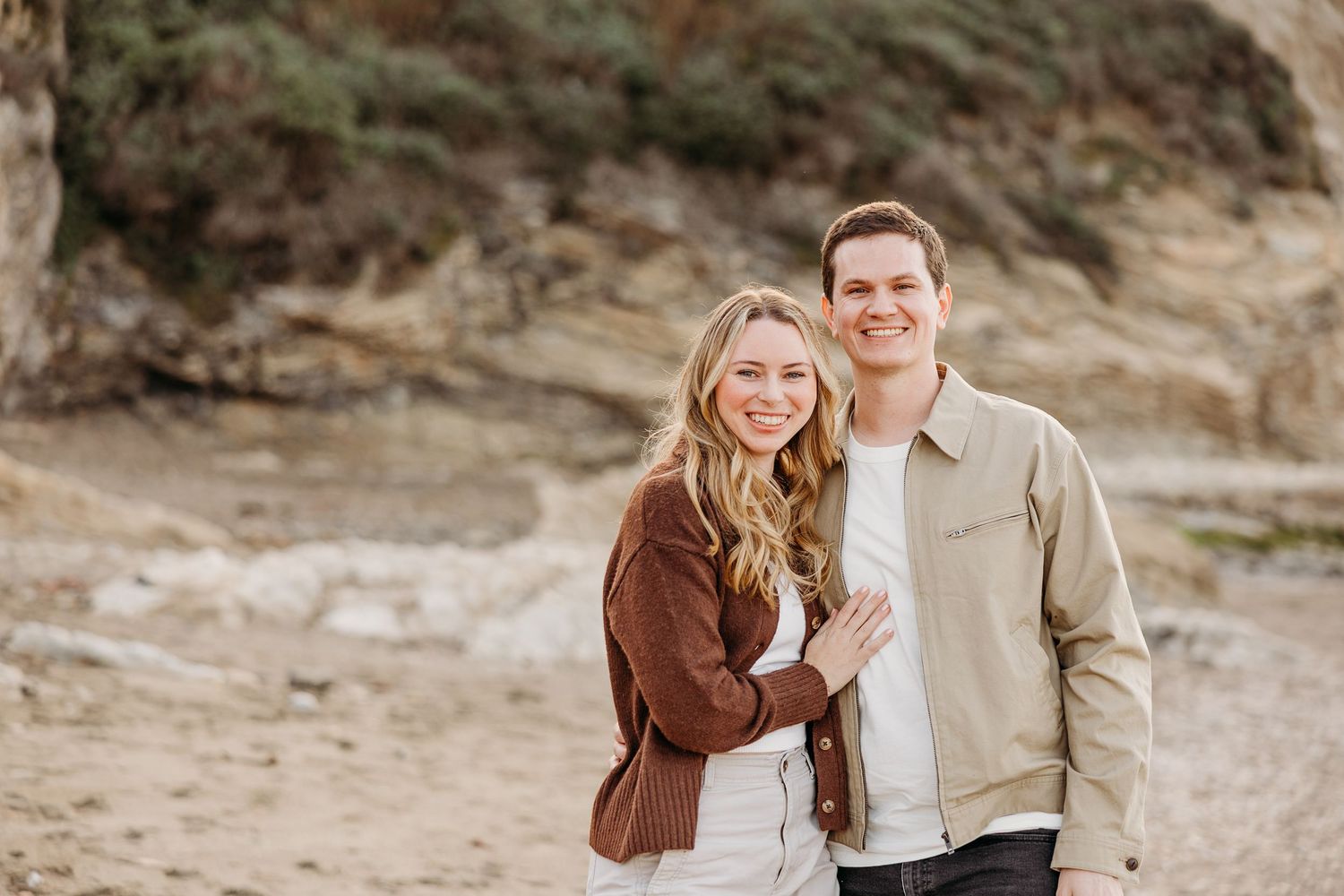 A couple poses together on a coastal path with rugged cliffs and natural scenery behind them.