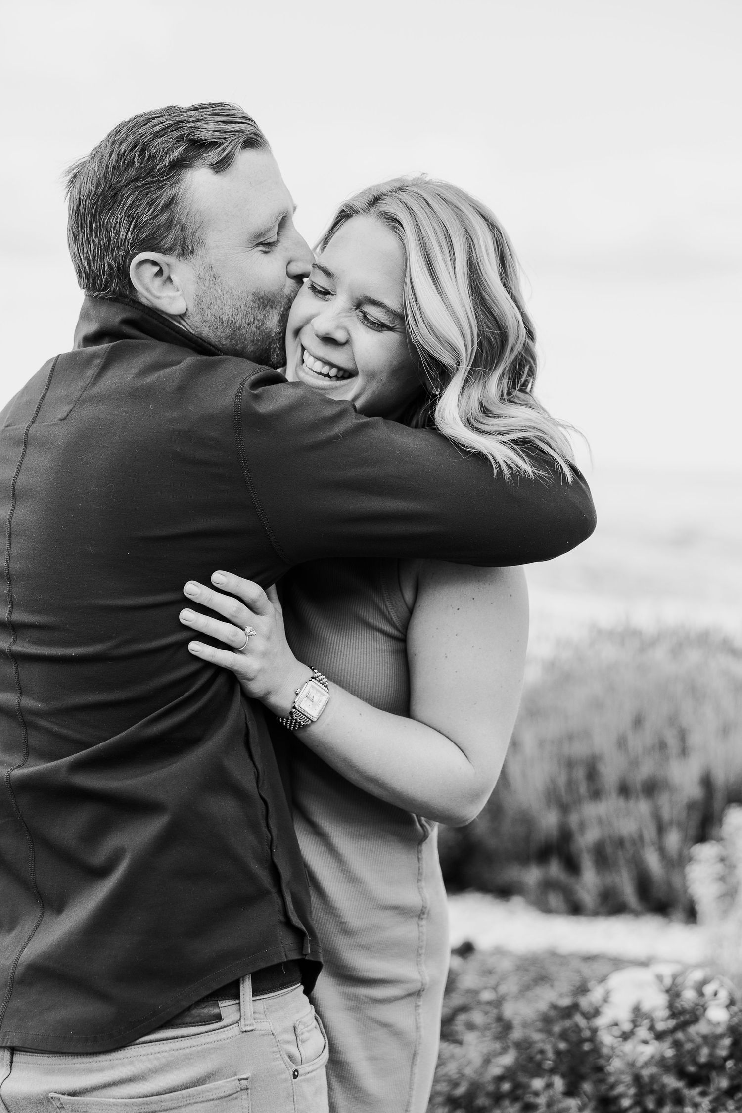 A couple embraces lovingly against a scenic mountain backdrop in black and white.