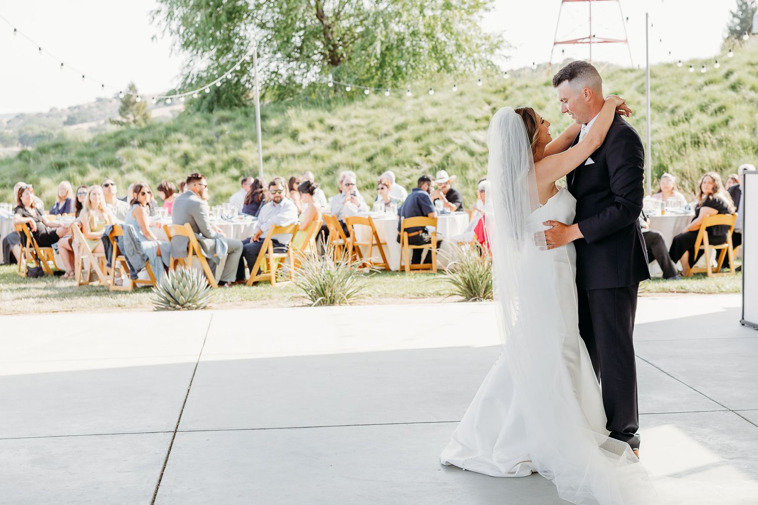 A couple shares their first dance during an outdoor wedding reception with guests seated at tables nearby.