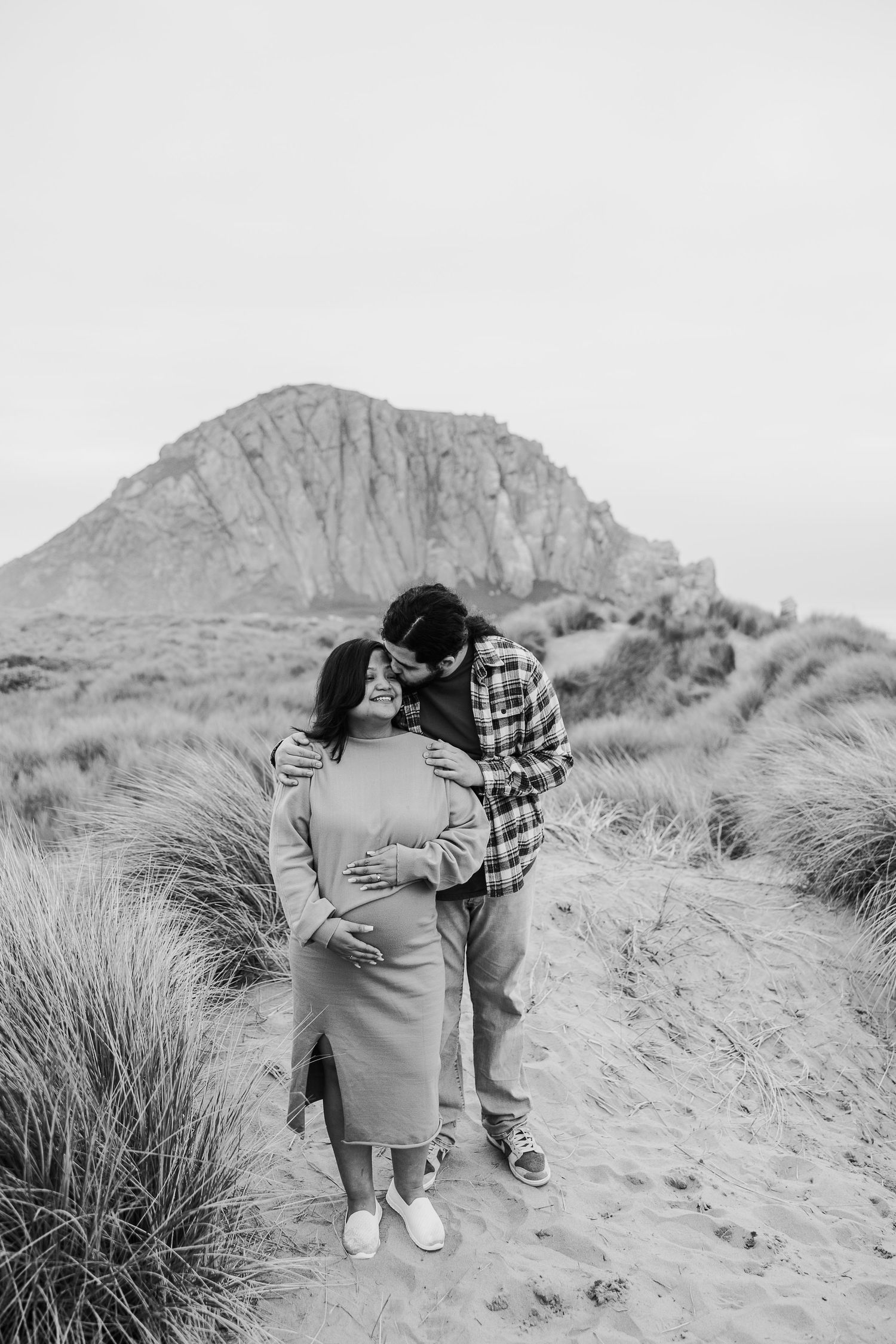 A romantic black and white portrait of a couple embracing on a sandy path with a mountain vista.