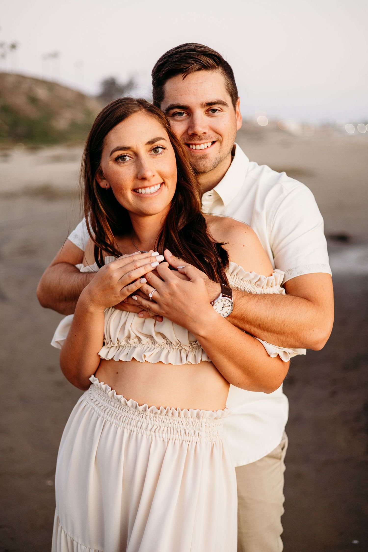 A couple shares an intimate moment in coordinated white outfits on a beach at sunset.