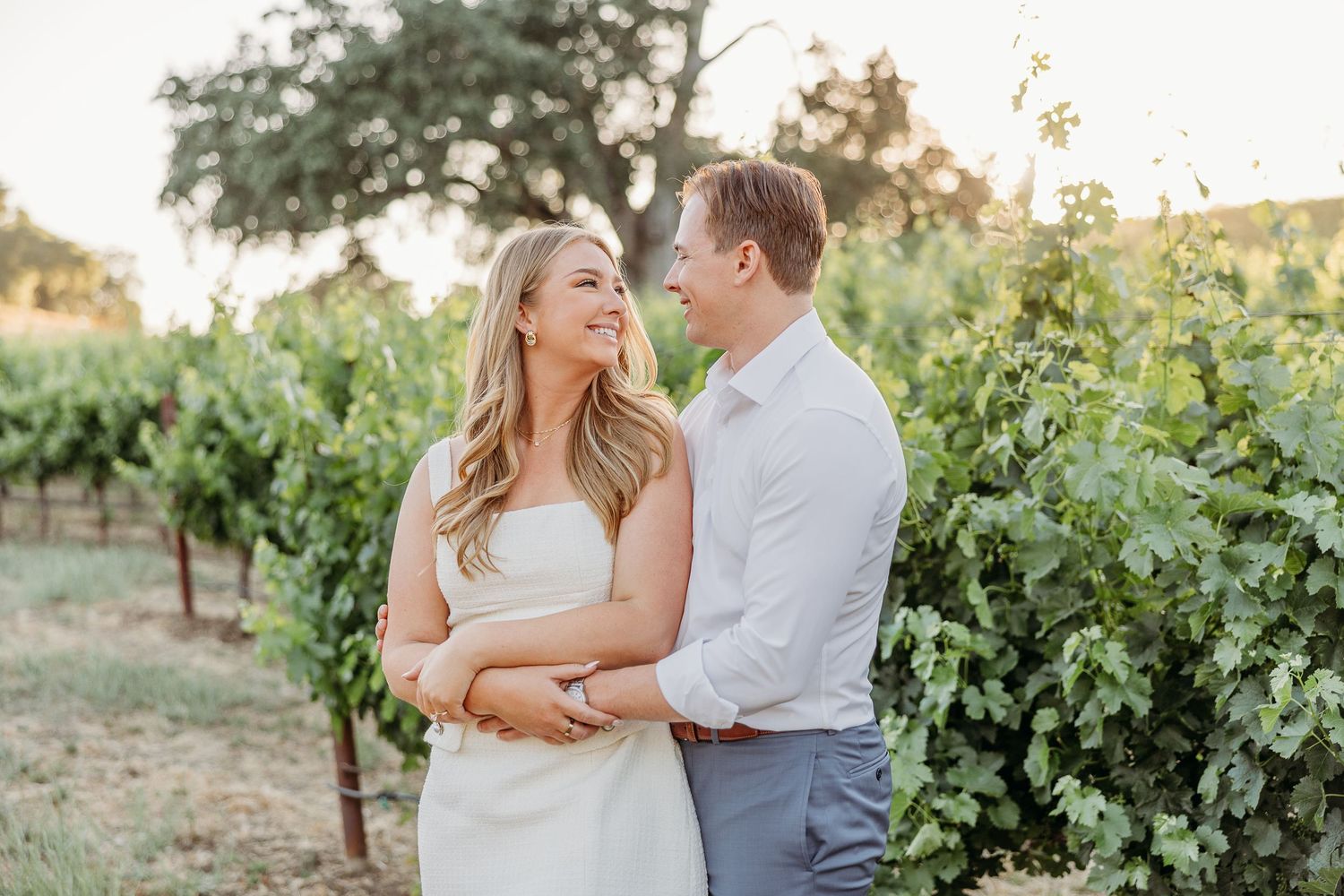 A couple shares a joyful moment together while standing in a sunlit vineyard at golden hour.