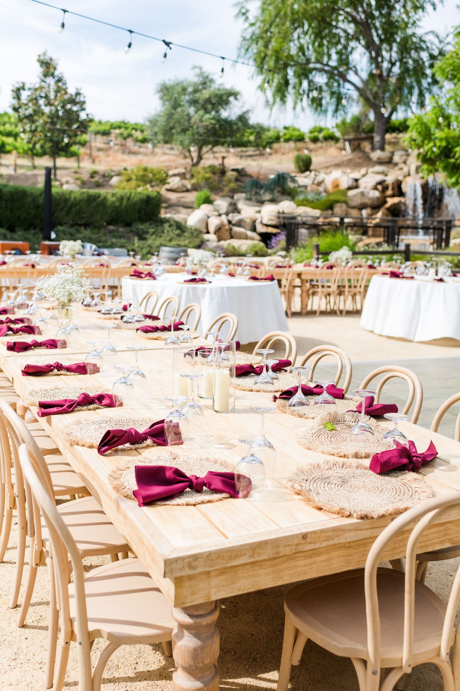 Elegant outdoor wedding reception table with burgundy accents and natural wooden chairs.