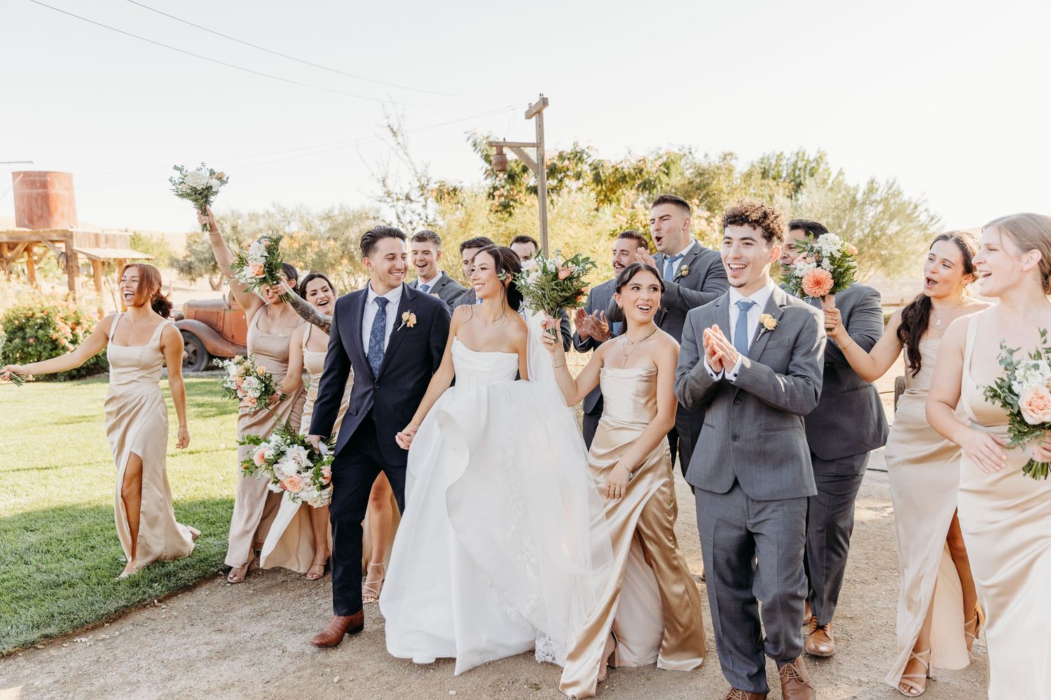 Wedding party members in gray and blush tones pose together on a sunny outdoor lawn.