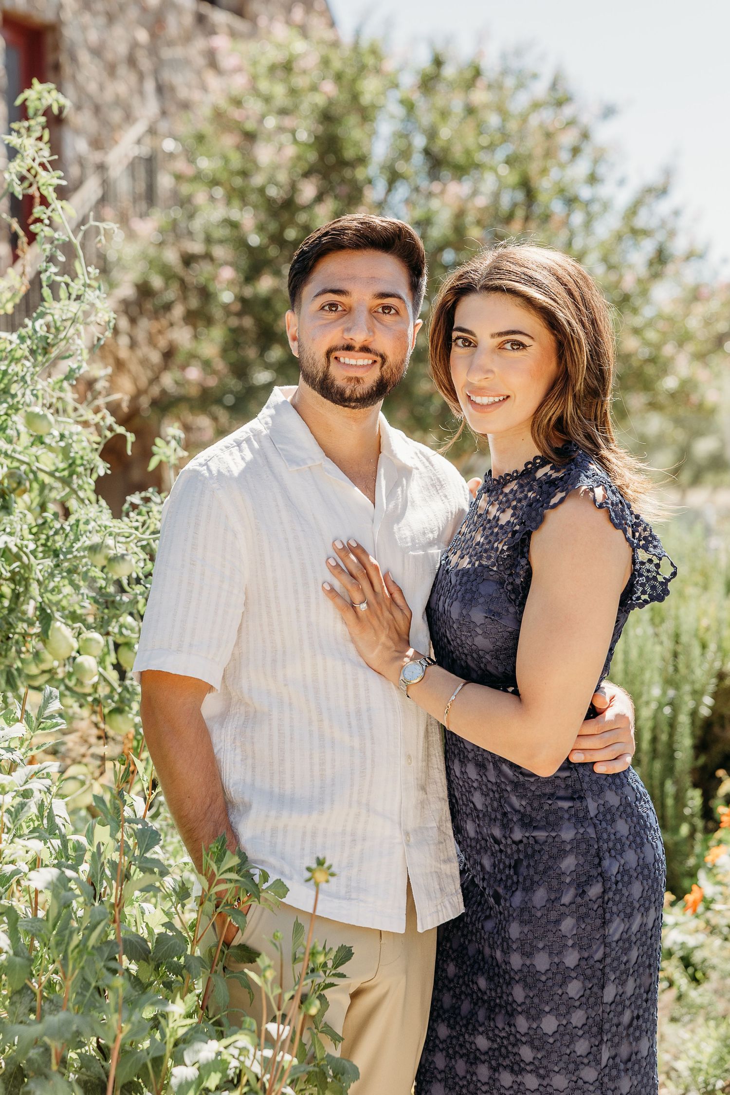 A couple poses together outdoors in a garden setting with blooming plants and greenery on a sunny day.