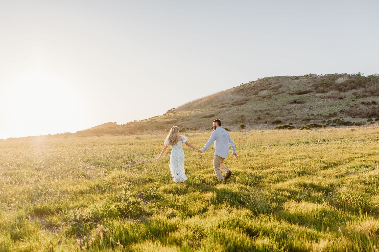 A group of people hold hands while walking through a sunlit meadow on a golden hillside at sunset.