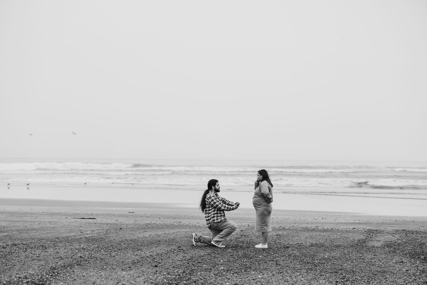 Black and white photo of a marriage proposal taking place on a misty beach with ocean waves in the background.